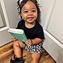 Amiri joined the competition — help win amazing prizes! child, toddler, smile, pigtails, bows, book, black_shirt, floral_shorts, sitting, wooden_floor, indoors, happy, cute, person, young_child, curly_hair, black_shoes, white_doors, holding_object, portrait