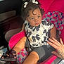 toddler, child, car_seat, pink, bows, curly_hair, black_sandals, black_shorts, patterned_shirt, tattooed_hand, indoor, seatbelt, car_interior, cute, portrait, sitting, young_child, person, footwear, expression