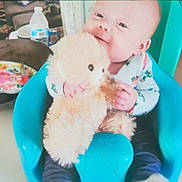 Jocelyn is registered to the contest to win money with this photo: baby, teddy_bear, chair, indoor, child, toy, water_bottle, plate, clothing, person, cute, happy, smile, seated, comfort, blue, furniture, infant, cozy, casual