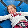 child, trampoline, blue, white_shirt, red_text, plaid_pants, young_child, outdoor, relaxed, lying_down, casual_clothing, portrait, smiling, happy, face, person, leisure, playtime, childhood, daylight