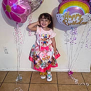 Gabriella is registered to the contest to win money with this photo: child, girl, birthday, balloons, pink, rainbow, smile, celebration, party, tiled_floor, wall, slippers, dress, cartoon_characters, happy, indoor, young, person, standing, festive