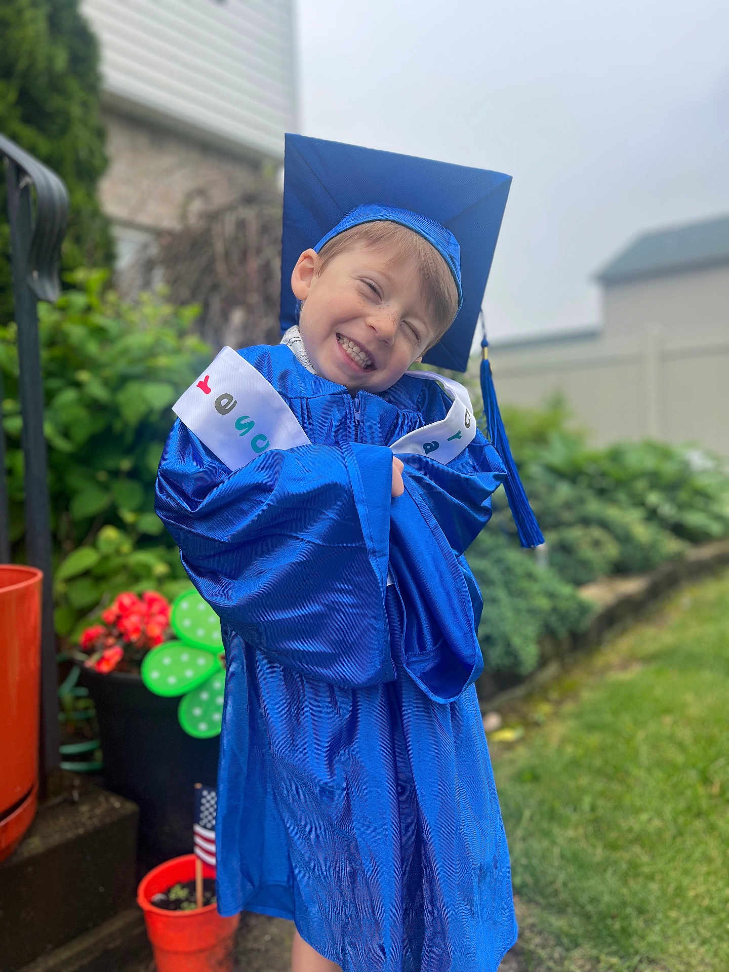 Joshua is registered to the contest to win money with this photo: child, boy, smiling, graduation_gown, graduation_cap, outdoor, garden, plants, flowers, greenery, happy, celebration, portrait, young, cute, blue, clothing, person, pot, american_flag
