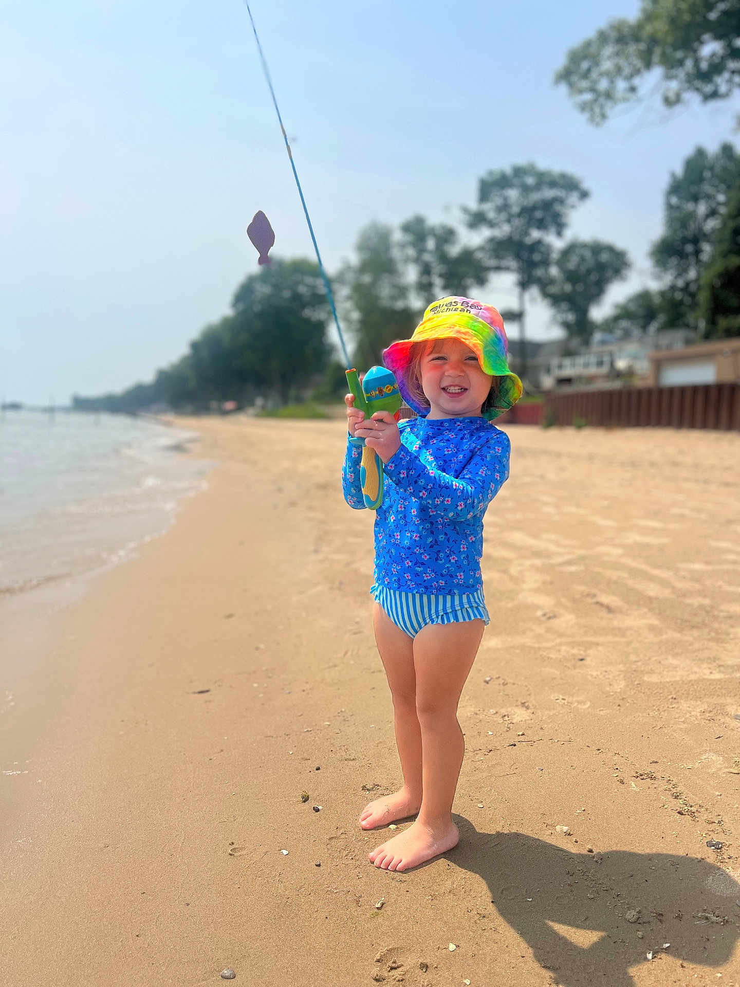 Avery is registered to the contest to win money with this photo: child, beach, sand, fishing_rod, hat, rainbow, smile, sunny, water, trees, blue_shirt, striped_swim_bottoms, barefoot, outdoor, summer, vacation, playful, nature, person, happy