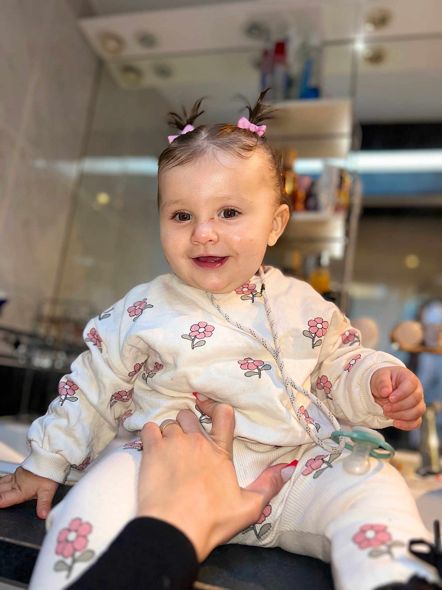 Catalya a rejoint le concours — aidez-le/la à gagner de superbes lots ! baby, child, smile, happy, pigtails, pink_bows, floral_outfit, sweatshirt, pants, pacifier, hand, adult_hand, indoor, mirror, bathroom, sink, blurry_background, cute, portrait, person