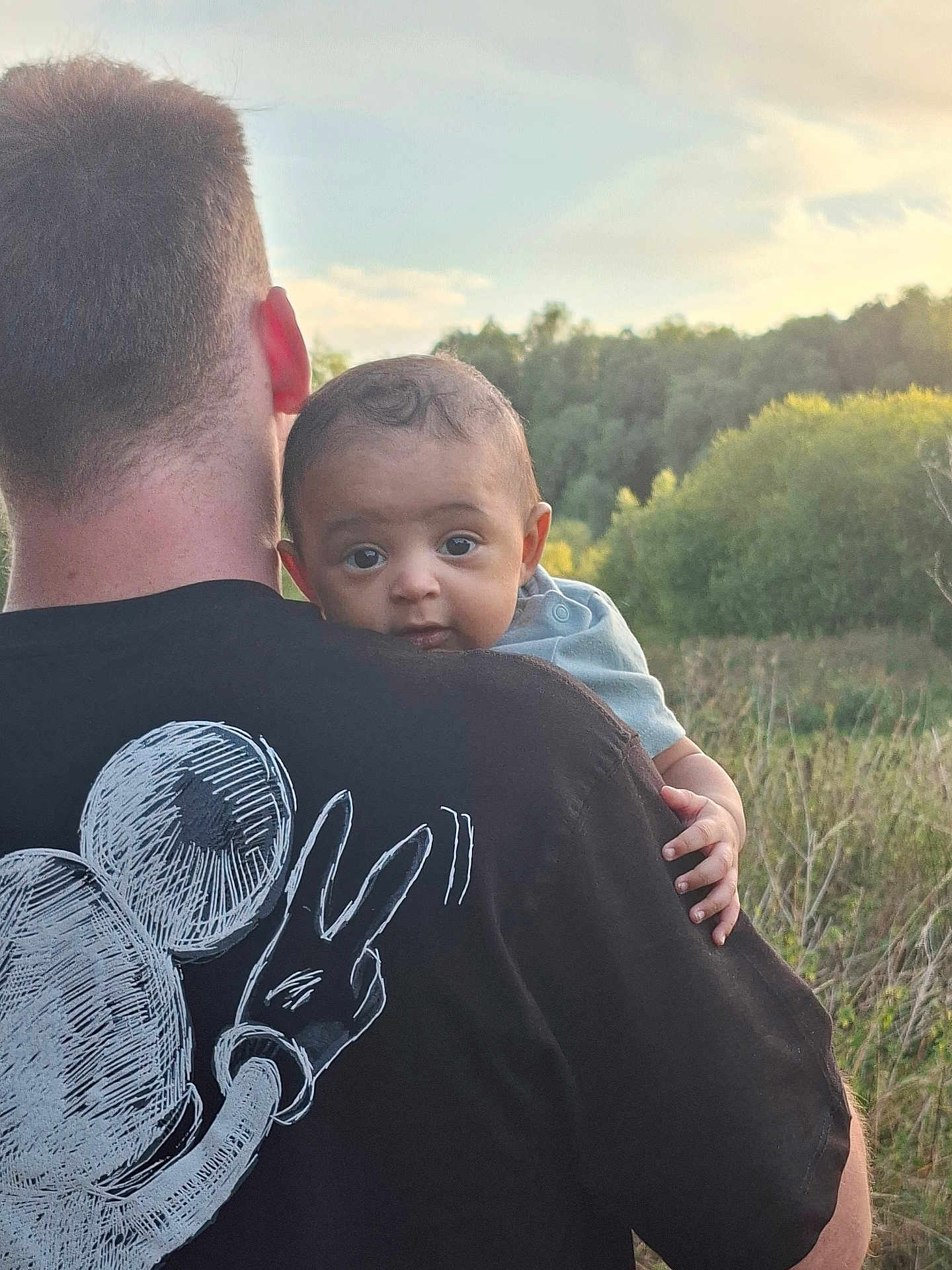 Ezra is registered to the contest to win money with this photo: baby, child, adult, back_view, mickey_mouse, black_shirt, peace_sign, outdoor, nature, greenery, sky, curious, portrait, holding, head, hand, short_hair, casual_clothing, daylight, family