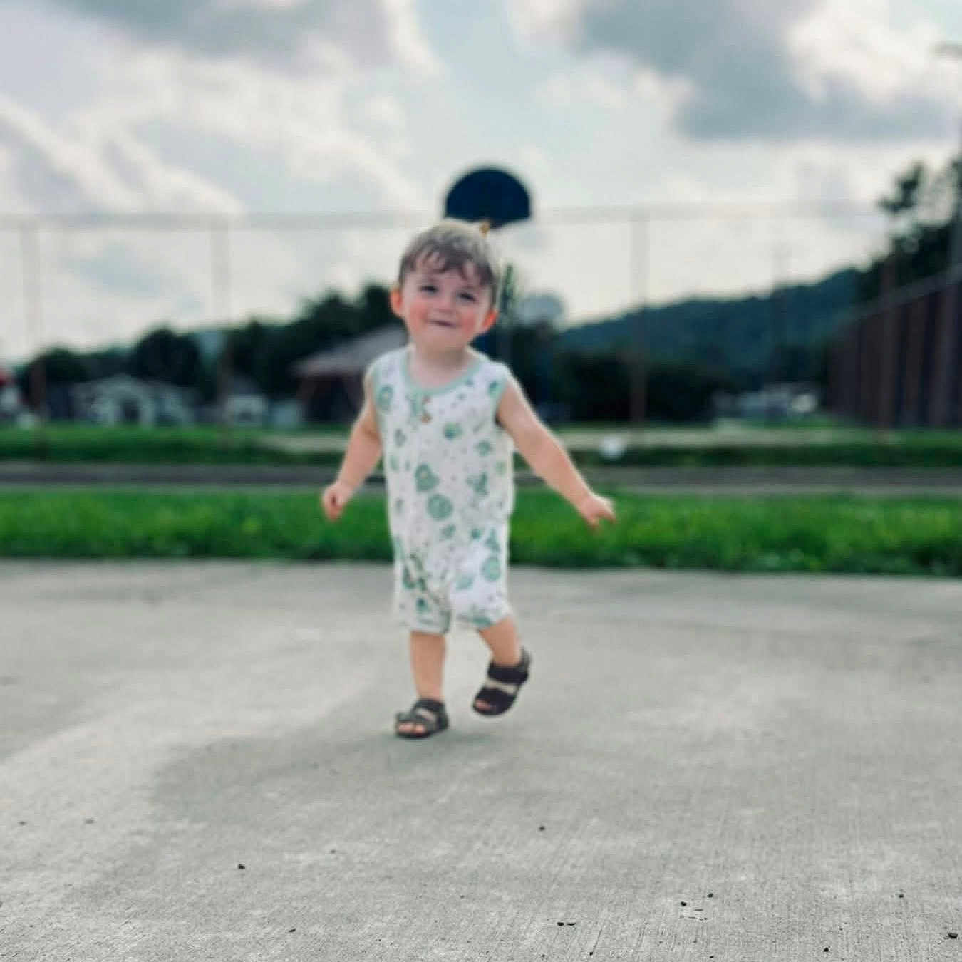 Zaydon joined the competition — help win amazing prizes! basketball_hoop, blurred_background, casual_clothing, child, clouds, concrete, daytime, fence, grass, greenery, outdoor, person, playground, romper, sandals, sky, summer, toddler, walking, young_child
