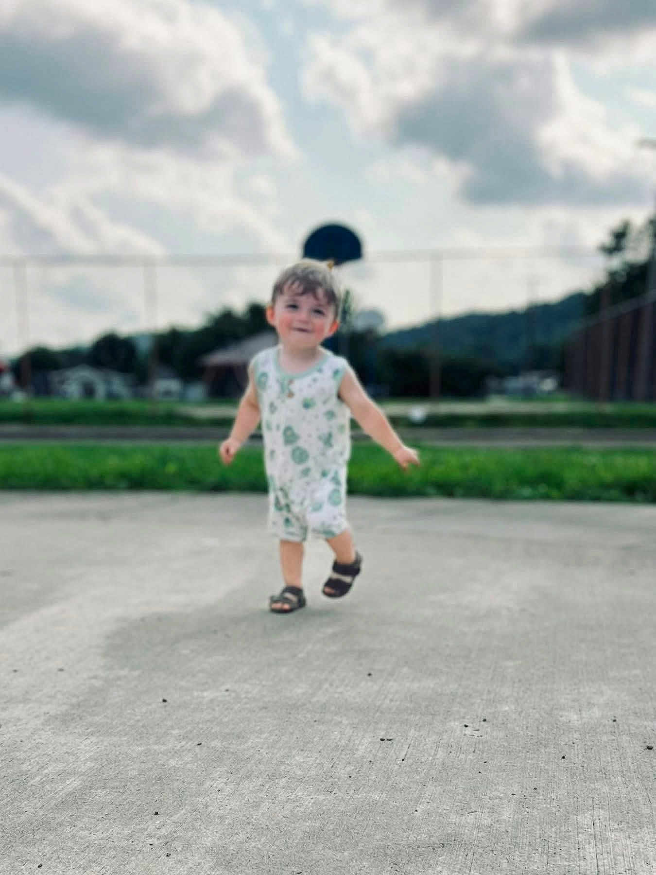 Zaydon joined the competition — help win amazing prizes! toddler, child, outdoor, concrete, basketball_hoop, grass, clouds, sky, greenery, romper, sandals, walking, person, young_child, daytime, fence, blurred_background, playground, summer, casual_clothing