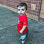 toddler, child, boy, red_shirt, nike, shorts, socks, bear_socks, sidewalk, brick_wall, outdoor, curious, standing, casual_clothing, young_child, portrait, concrete, urban, daylight, cute