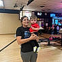 Zaydon is registered to the contest to win money with this photo: woman, toddler, bowling_alley, bowling_balls, people, indoor, casual_clothing, smile, holding, child, floor, screen, furniture, background, activity, recreation, sport, family, happy, interaction