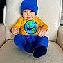 baby, child, blue_beanie, yellow_shirt, globe_design, smiley_face, pants, socks, chair, soft_texture, indoor, portrait, cute, happy, seated, infant, clothing, face, hands, feet
