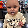 Kalani is registered to the contest to win money with this photo: baby, child, face, messy_face, cream, clothing, shirt, text_on_clothing, indoor, store, table, chair, person, curly_hair, cute, closeup, sitting, expression, large_eyes, hand