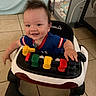 baby, child, smiling, happy, portrait, indoor, walker, activity_center, toy, infant, blue_shirt, short_sleeves, seated, playtime, tile_floor, home, furniture, baby_gear, colorful, close_up