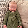 baby, child, smile, green_onesie, couch, cushion, indoor, portrait, happy, infant, person, face, seated, cozy, home, fabric, texture, cute, young, relaxed