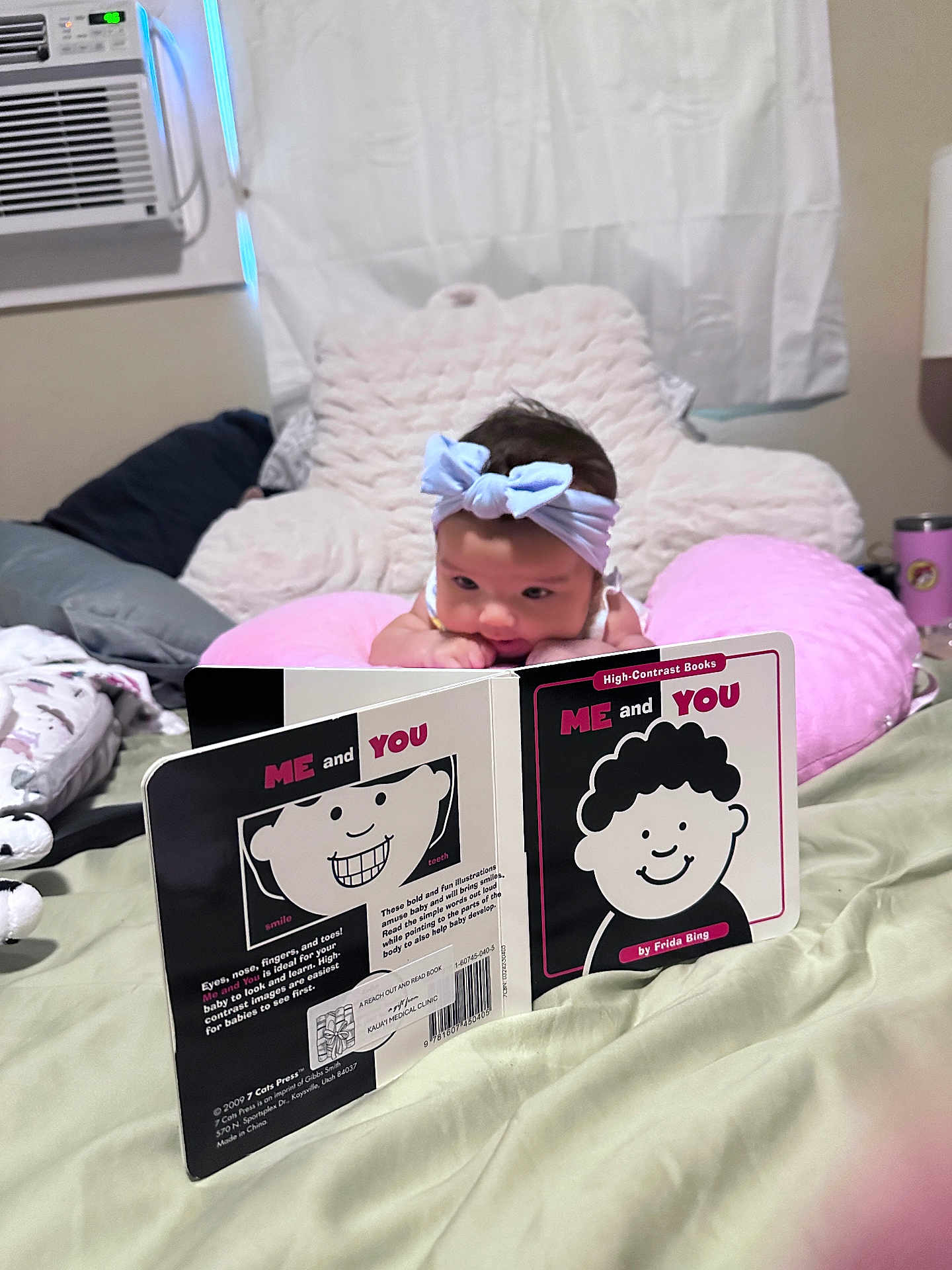 Magnolia is registered to the contest to win money with this photo: baby, headband, pink_pillow, book, high_contrast_book, bed, blanket, indoor, air_conditioner, curtains, reading, infant, child, face, person, soft_toys, cozy, focus, portrait, cute