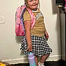 Layla joined the competition — help win amazing prizes! child, girl, backpack, water_bottle, indoor, plaid_skirt, sneakers, white_socks, standing, smiling, casual_clothing, person, floor, wall, cabinet, hair_tied_back, young, ready_for_school, accessories, portrait