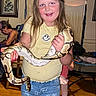 Layla is registered to the contest to win money with this photo: child, girl, snake, animal, indoor, person, smiling, jeans, yellow_shirt, wooden_floor, furniture, adult, family, painting, home, long_hair, casual_clothing, holding, reptile, living_room