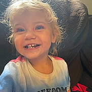 Victoria joined the competition — help win amazing prizes! child, toddler, smiling, happy, portrait, face, blonde_hair, curly_hair, gap_tooth, teeth, indoor, couch, home, sunlight, backlit, colorful_clothing, t_shirt, freedom_text, close_up, joyful