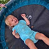child, toddler, laughing, smiling, blond_hair, blue_outfit, trampoline, outdoor, grass, garden, playground, recreation, lying_down, raised_arm, happy, portrait, closeup, daylight, leaf, playful