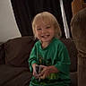 Victoria is registered to the contest to win money with this photo: child, toddler, smiling, happy, portrait, indoor, couch, seated, controller, videogame, green_shirt, blonde_hair, blue_eyes, barefoot, curtains, living_room, furniture, playful, home, photograph