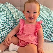 Charlotte is registered to the contest to win money with this photo: baby, child, smiling, pink_dress, sitting, pillow, bed, happy, infant, cute, indoors, person, face, blue_eyes, sock, furniture, pattern, home, young_child, portrait