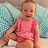 baby, child, smiling, pink_dress, sitting, pillow, bed, happy, infant, cute, indoors, person, face, blue_eyes, sock, furniture, pattern, home, young_child, portrait