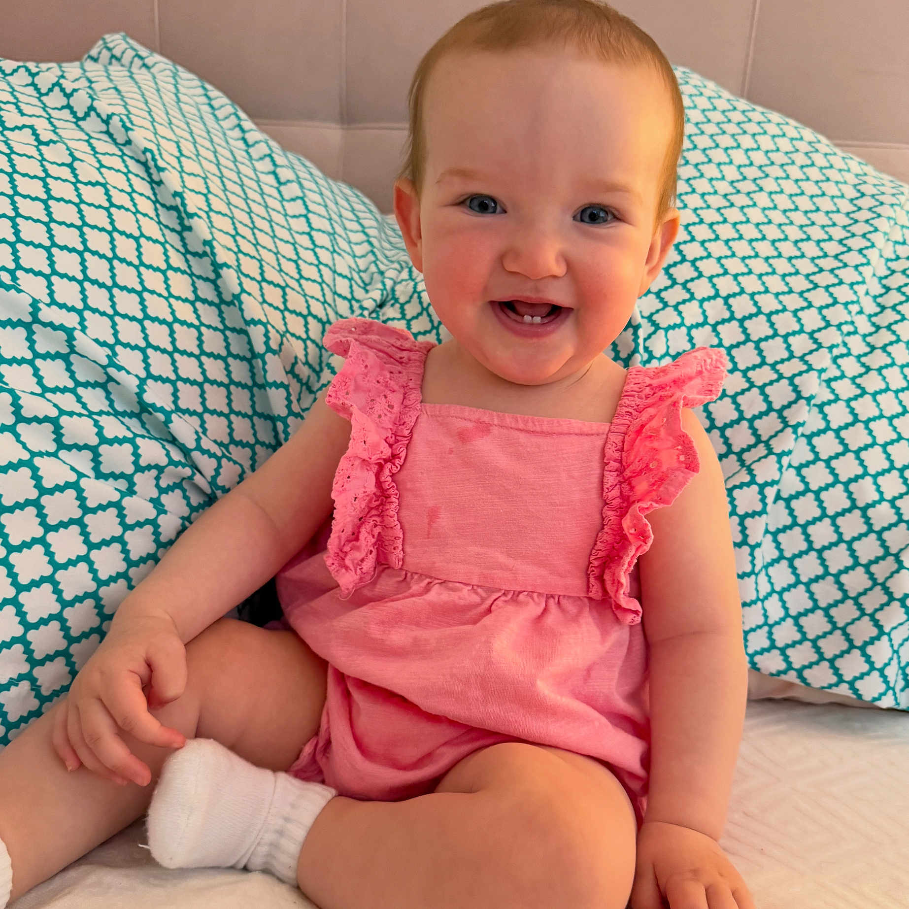 Charlotte is registered to the contest to win money with this photo: baby, child, smiling, pink_dress, sitting, pillow, bed, happy, infant, cute, indoors, person, face, blue_eyes, sock, furniture, pattern, home, young_child, portrait