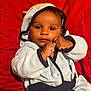 baby, child, infant, face, eyes, hands, hood, robe, clothing, fist, cute, portrait, red_background, smiling, skin, person, sitting, indoor, young, expression