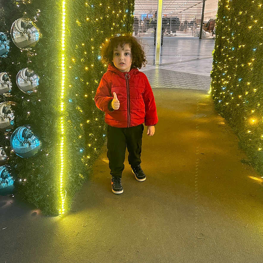 Nidhal participe au concours pour gagner de l'argent avec cette photo : black_pants, casual_clothing, child, curly_hair, festive_lights, floor, holiday_decor, indoor, modern, person, photography, red_jacket, reflection, shopping_mall, silver_ornaments, smiling, sneakers, thumbs_up, tunnel, yellow_lights