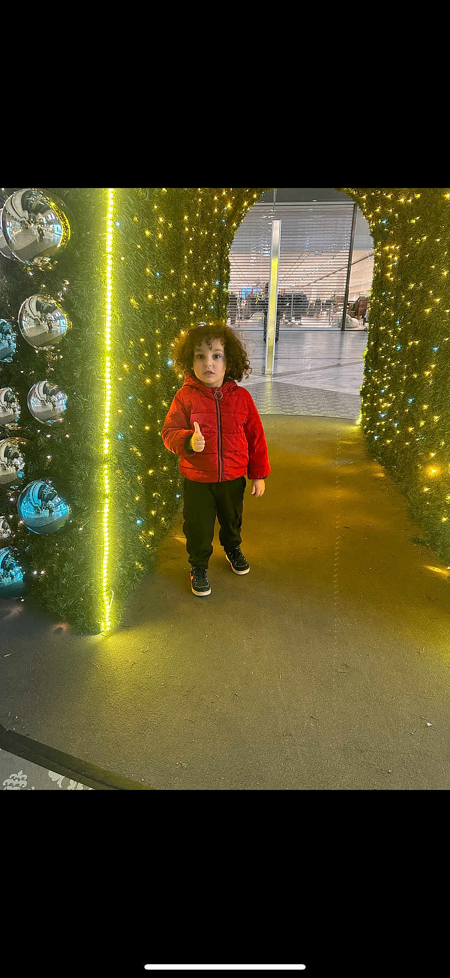 Nidhal participe au concours pour gagner de l'argent avec cette photo : child, curly_hair, red_jacket, thumbs_up, festive_lights, yellow_lights, silver_ornaments, tunnel, indoor, shopping_mall, floor, casual_clothing, black_pants, sneakers, holiday_decor, smiling, person, photography, modern, reflection