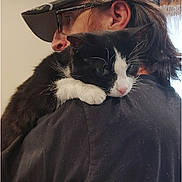 Dylan is registered to the contest to win money with this photo: person, cat, black_and_white_cat, cap, glasses, indoor, hugging, shoulder, wall, wooden_beam, window, curtain, dark_shirt, long_hair, side_view, pet, animal, comfort, affection, casual