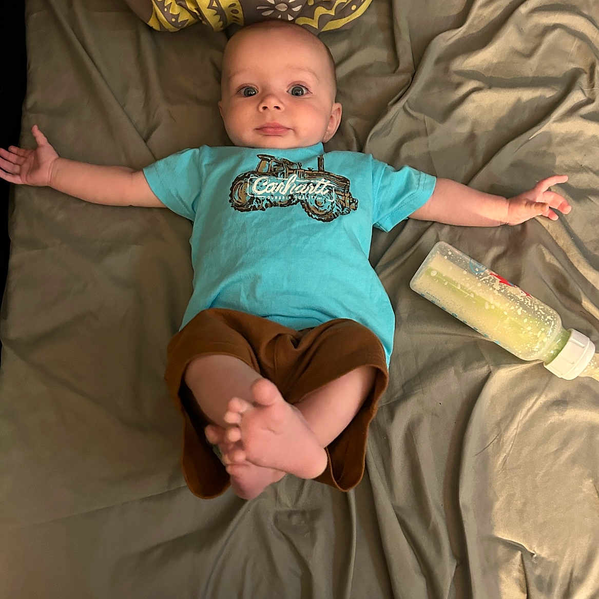 Kashton joined the competition — help win amazing prizes! arms, baby, bed, blanket, blue_shirt, bottle, brown_pants, clothing, comfort, curious, cute, expression, fabric, face, feet, indoor, infant, lying_down, person, wood_floor