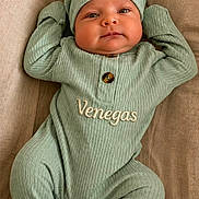 Ezekiel joined the competition — help win amazing prizes! baby, infant, child, green_outfit, hat, personalized_clothing, embroidered_text, lying_down, beige_background, cute, face, eyes, smile, hands_up, footed_pajamas, cozy, portrait, newborn, soft_lighting, indoor