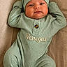 baby, infant, child, green_outfit, hat, personalized_clothing, embroidered_text, lying_down, beige_background, cute, face, eyes, smile, hands_up, footed_pajamas, cozy, portrait, newborn, soft_lighting, indoor