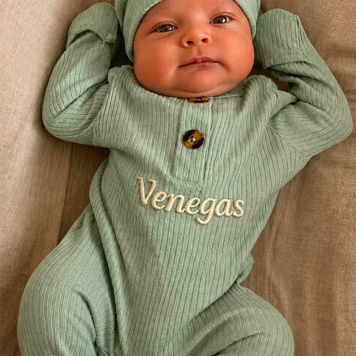Ezekiel joined the competition — help win amazing prizes! baby, infant, child, green_outfit, hat, personalized_clothing, embroidered_text, lying_down, beige_background, cute, face, eyes, smile, hands_up, footed_pajamas, cozy, portrait, newborn, soft_lighting, indoor