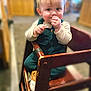 Avery is registered to the contest to win money with this photo: baby, child, high_chair, wooden_chair, indoor, curly_hair, clothing, footwear, smile, face, person, seated, cute, young_child, infant, hand, furniture, blurry_background, expression, eating