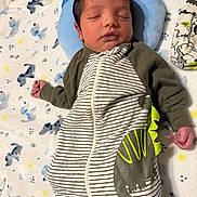 Jason is registered to the contest to win money with this photo: baby, newborn, sleeping, infant, onesie, striped_clothing, dinosaur_design, blanket, pillow, patterned_blanket, cozy, peaceful, resting, child, indoor, soft_texture, cute, person, hand, head