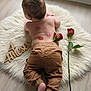 baby, infant, back_view, brown_pants, red_rose, rose_petals, fluffy_rug, wooden_sign, floor, skin, natural_light, soft_texture, lying_down, baby_skin, cute, indoors, child, newborn, age_marker, decor