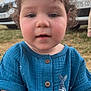 toddler, child, face, curly_hair, blue_shirt, buttons, outdoor, grass, car, vehicle, license_plate, daylight, portrait, young_child, casual_clothing, person, closeup, expressionless, nature, sitting