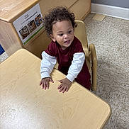 Maurice joined the competition — help win amazing prizes! child, toddler, curly_hair, maroon_clothing, white_sleeves, table, chair, classroom, floor_tiles, bulletin_board, wooden_furniture, indoor, person, playful_expression, young_child, sitting, hand_on_table, educational_environment, light_wood, curious