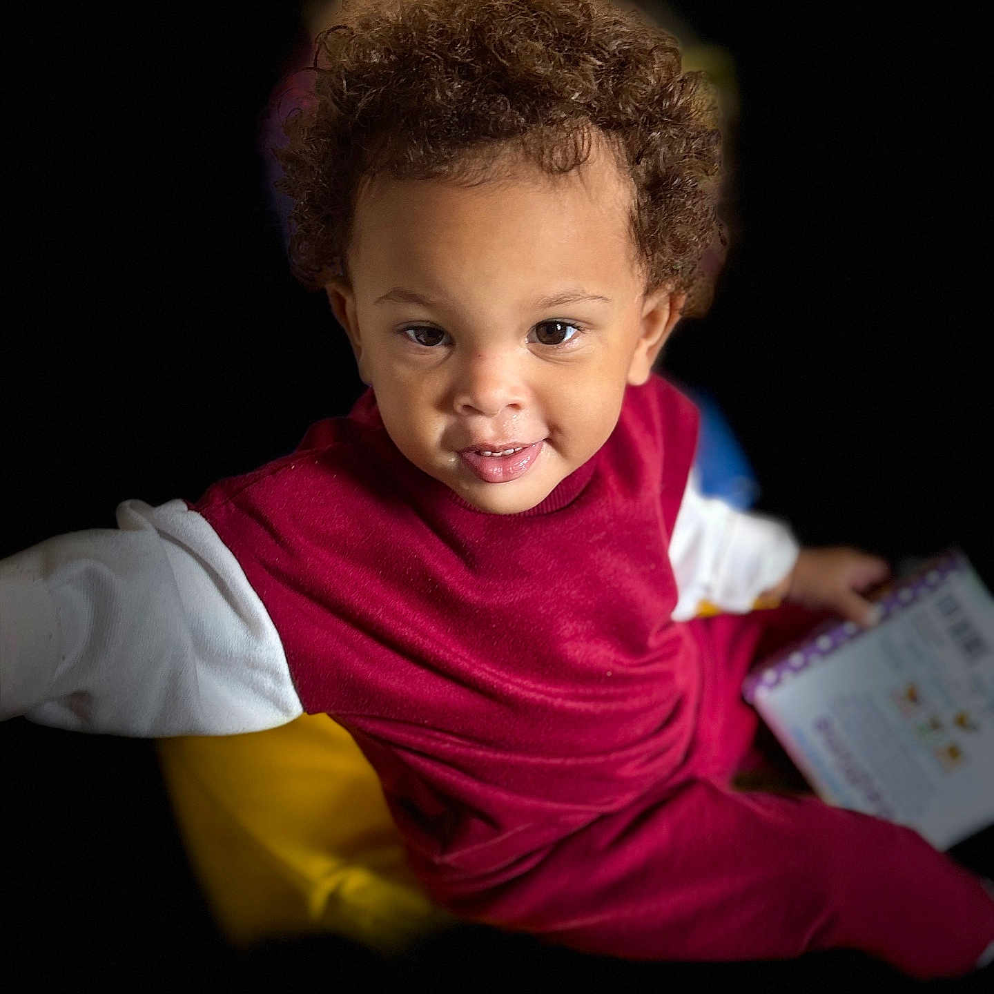 Maurice is registered to the contest to win money with this photo: child, toddler, curly_hair, red_clothing, white_sleeve, book, reading, smiling, portrait, indoor, dark_background, cute, young_child, person, happy, baby, seated, looking_up, playful, bright_colors