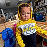 Maurice is registered to the contest to win money with this photo: toddler, child, yellow_hoodie, indoor, playroom, toy, rocking_horse, blue_toy, serious_expression, person, curly_hair, wooden_furniture, colorful, floor, window_blinds, air_conditioner, educational_materials, wall_decor, daylight, casual_clothing
