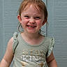 child, girl, smiling, happy, portrait, close_up, face, teeth, eyes, hair, ponytail, shirt, clothing, kindness, indoor, blue_wall, standing, playful, funny_expression, photography