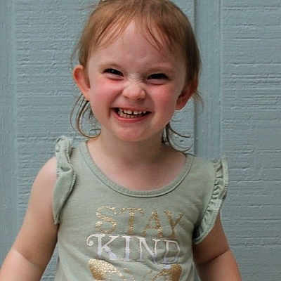 Paysen is registered to the contest to win money with this photo: child, girl, smiling, happy, portrait, close_up, face, teeth, eyes, hair, ponytail, shirt, clothing, kindness, indoor, blue_wall, standing, playful, funny_expression, photography