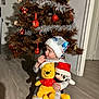 child, toddler, plush_toy, winnie_the_pooh, mickey_mouse, christmas_tree, christmas_ornaments, holiday, hat, floor, indoor, decorations, toy, festive, santa_hat, curious, sitting, winter_clothing, celebration, cute