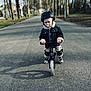 balance_bike, casual_clothing, child, daytime, helmet, jacket, knee_pads, outdoor, pacifier, park, person, protective_gear, road, safety, shoes, sidewalk, street, toddler, trees, young_child