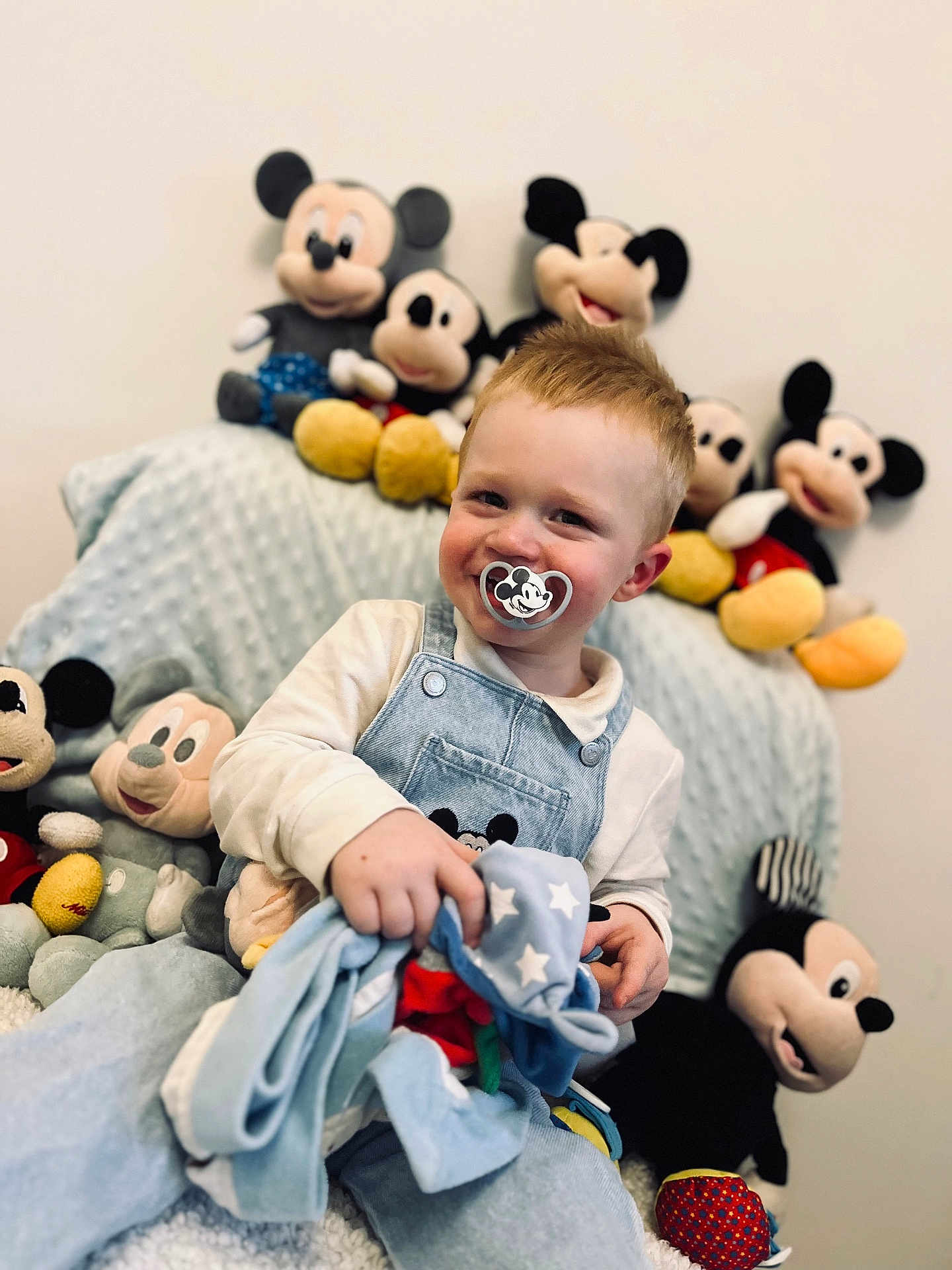 Elio participe au concours pour gagner de l'argent avec cette photo : toddler, child, pacifier, mickey_mouse, plush_toys, stuffed_animals, blanket, denim_overalls, smile, happy, indoor, cozy, seated, cute, toy_collection, soft_toys, person, face, playful, casual_clothing