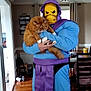 Kylo joined the competition — help win amazing prizes! person, costume, skeletor, cat, orange_cat, fluffy_cat, indoor, wooden_floor, curtains, chair, table, boots, purple, blue, mask, holding, pet, fur, home, funny