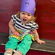 Olivia is registered to the contest to win money with this photo: toddler, child, hat, paper_craft, turkey, purple, striped_shirt, colorful_clothing, sitting, bench, wooden_bench, red_background, shoes, smiling, happy, curly_hair, indoor, playful, cute, portrait