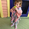 Phoebe is registered to the contest to win money with this photo: child, toddler, girl, apple, bite, dress, floral, jacket, braids, sandals, floor, wooden_floor, colorful_wall, pastel_colors, pink, yellow, purple, standing, outdoor, portrait