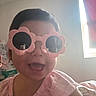 Marisol is registered to the contest to win money with this photo: toddler, child, pink_sunglasses, flower_shaped_glasses, smiling, happy, indoor, window, natural_light, pink_clothing, face, head, person, cute, young_child, baby, room, furniture, reflection, casual